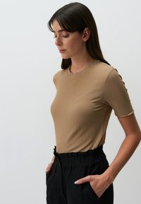 Lichtbruin geribd T-shirt met korte mouwen en een scalloped halslijn. Combineert met zwarte relaxed-fit broek, handen in de zakken. Soepele textuur.