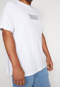 Camiseta de algodón blanca de manga corta con un logotipo gris de Calvin Klein en un parche rectangular, corte relajado y cuello redondo.