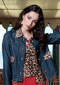 Jeune femme aux longs cheveux foncés portant une veste en jean par-dessus un haut à imprimé léopard et un pantalon rouge, posant à l'intérieur avec une expression neutre.