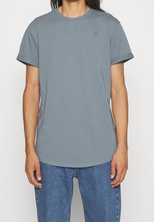 Person iført en ensfarvet blågrå kortærmet t-shirt og blå jeans, stående mod en ens hvid baggrund.