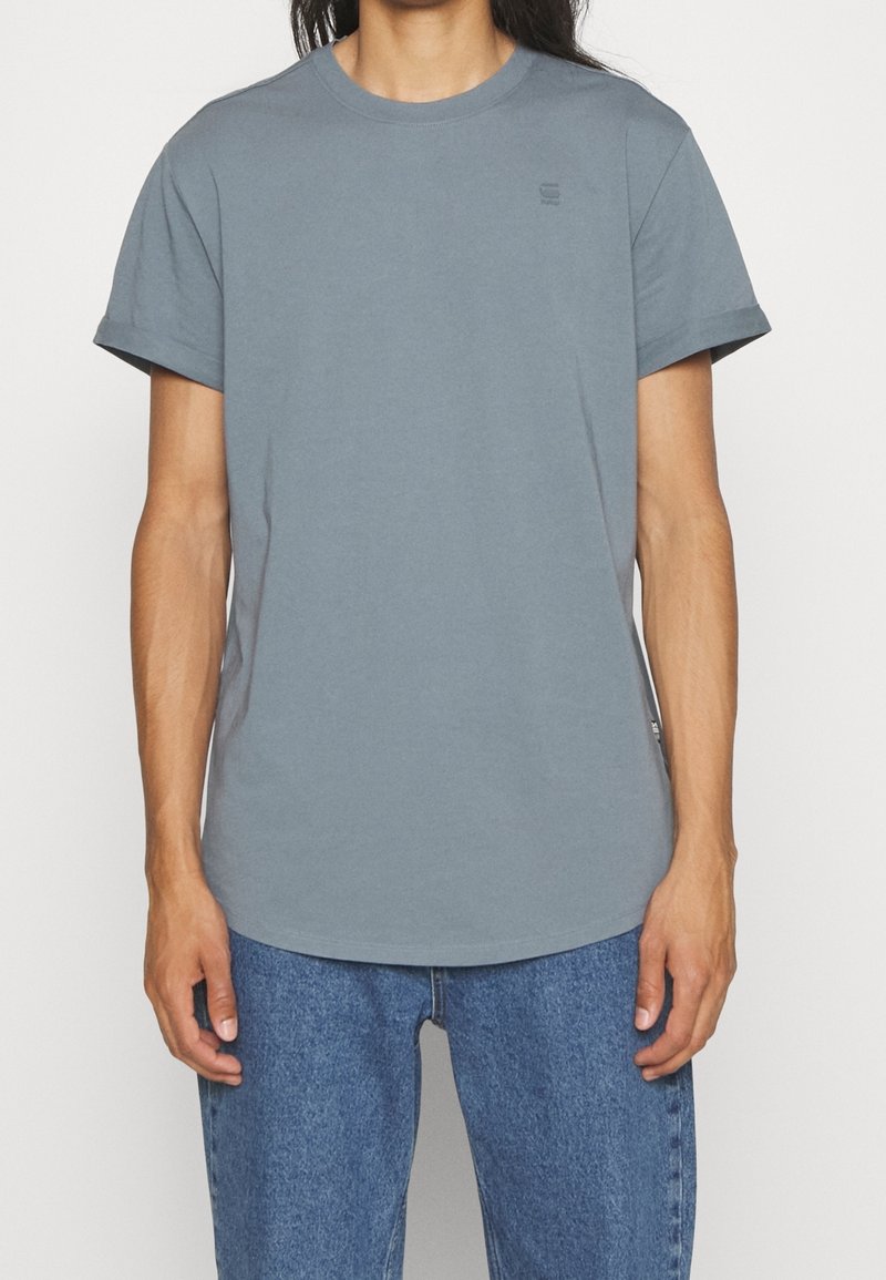 Personne portant un t-shirt bleu-gris uni à manches courtes et un jean bleu, debout devant un fond blanc uni.