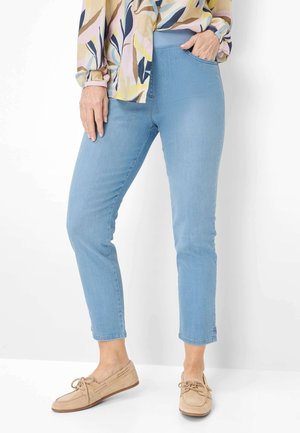 Vrouw draagt lichtblauwe, enkelhoge, strakke jeans, een kleurrijk gebloesd patroon blouse en beige mocassin schoenen.