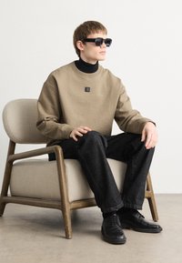 Bežna različica s kapuljačo z logotipom, črna turtleneck, temne kavbojke in črne čevlje. Sedi na svetli fotelji proti nevtralni ozadju. Sončna očala dopolnjujejo izgled.