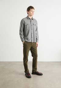 Chemise grise structurée avec deux poches poitrine, assortie à un pantalon vert olive et des chaussures noires avec des accents bordeaux. Design simple et moderne.