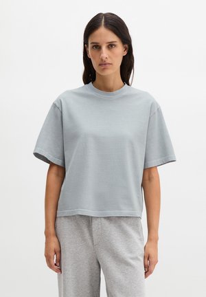 Kobieta o długich ciemnych włosach nosi luźny, jasnoszary top z krótkim rękawem i dopasowane szare spodnie na tle jednolitej, białej ściany.