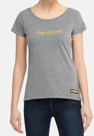 Kobieta nosząca dopasowany szary t-shirt z okrągłym dekoltem z żółtym napisem na piersi oraz ciemnoniebieskie dżinsy.