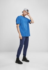 Tricou albastru din bumbac, cu mâneci scurte, combinat cu pantaloni de trening bleumarin și adidași negri. Pălărie gri cu clapetă pe cap.