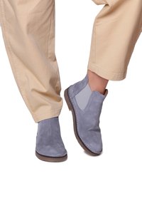 Pantalons beiges légèrement retroussés au-dessus des chevilles et pieds portant des bottines en daim grises avec des panneaux élastiques sur le côté et des semelles brunes.