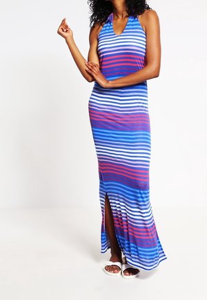 Kobieta w niebiesko-biało-czerwonej maxi sukience w poziome paski bez rękawów z rozcięciem z przodu i białych sandałach, stojąca na białym tle.