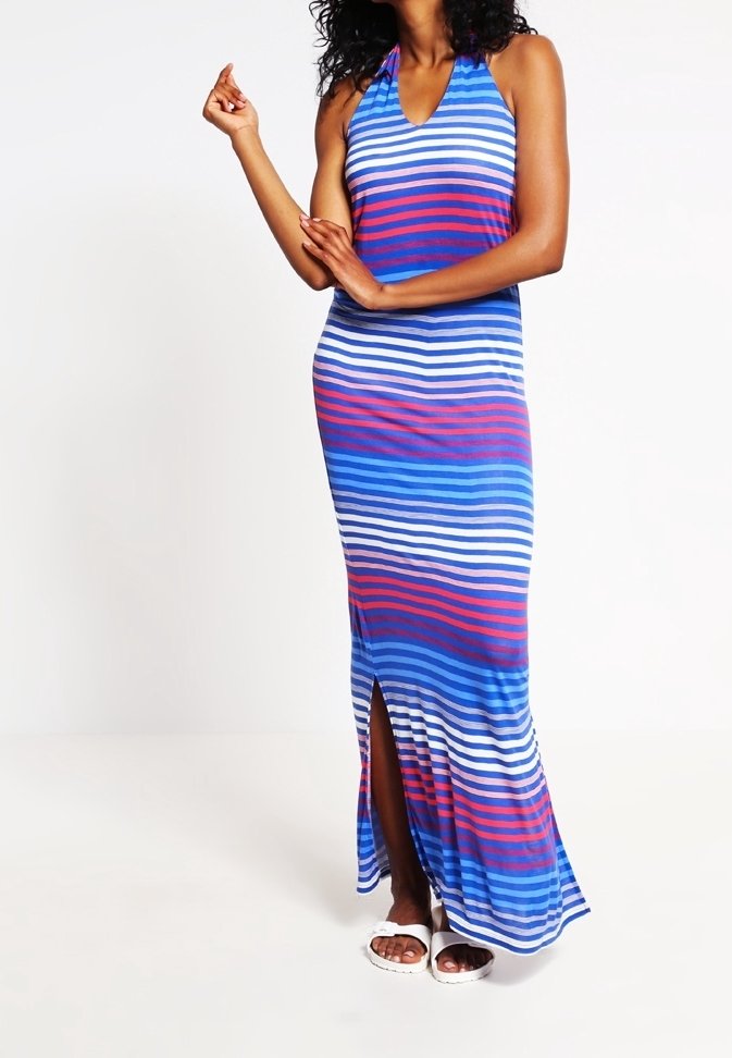 Femme portant une robe maxi sans manches à rayures horizontales bleues, blanches et rouges avec une fente avant et des sandales blanches, debout devant un fond blanc.