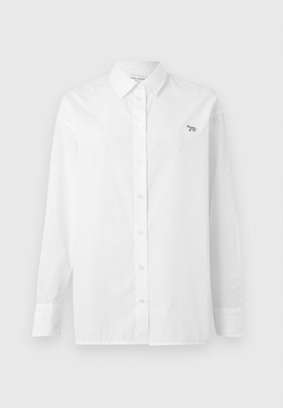 Biela košeľa s dlhým rukávom vyrobená z bavlny, so skladacím golierom, prednými knoflíkmi a malým vyšitým logom na ľavom prsnom vrecku.