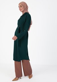 Long, dark green coat with a belt and button-up front, paired with brown wide-leg pants and light pink hijab. Smooth fabric texture.