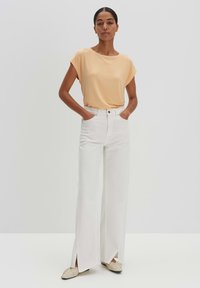 Lichtoranje korte mouwen top gecombineerd met hoge taille witte wijde broek met zijsplitten, aangevuld met beige platte schoenen.