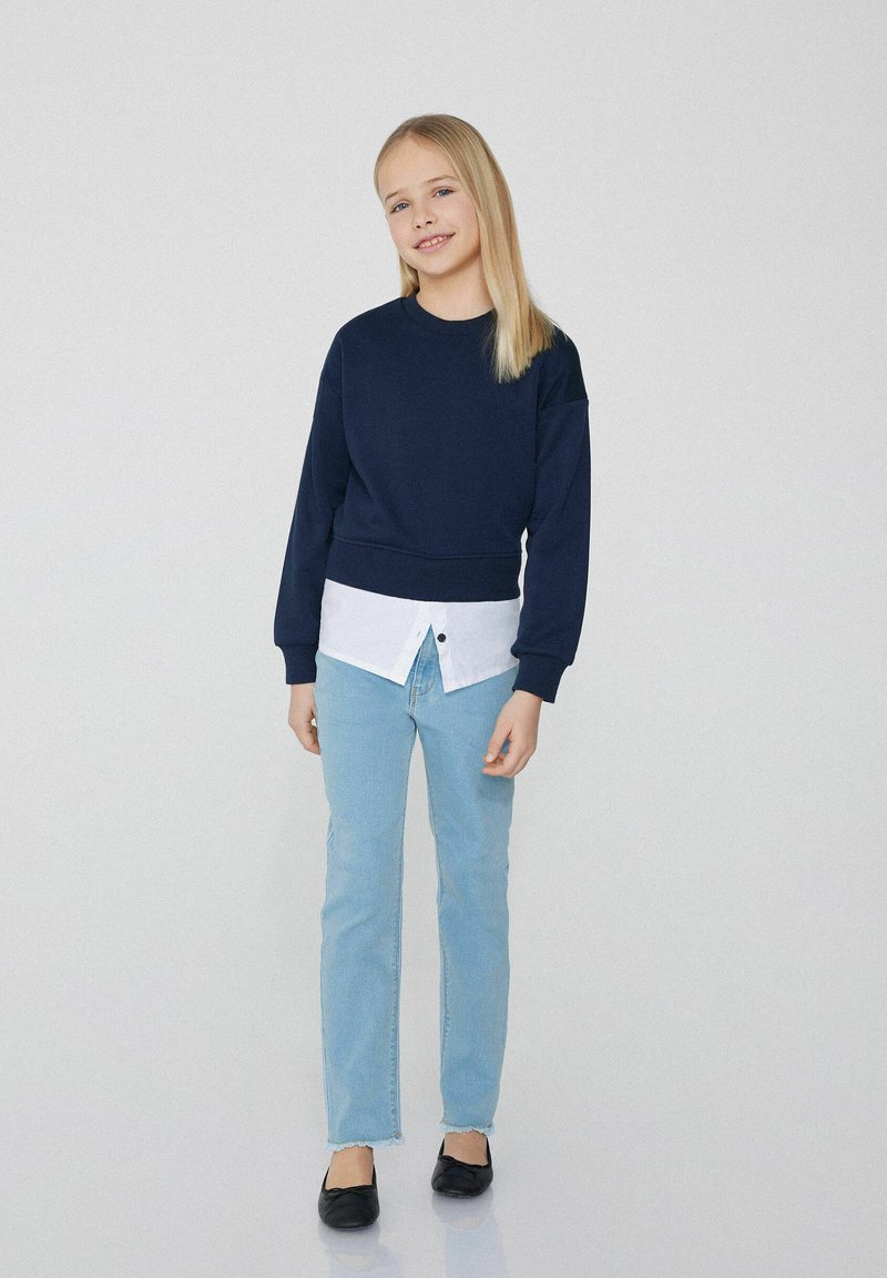 Námořnická mikina přes bílou košili s knoflíky, v kombinaci se světle modrými džíny a černými balerínami. Oblečení má ležérní vrstvený vzhled.