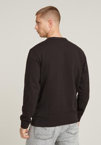Black sweatshirt with long sleeves and a crew neck, featuring a smooth texture and ribbed cuffs. The back view shows minimal design details.