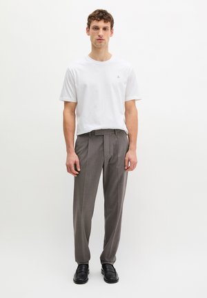 Jonge man die staat, gekleed in een wit T-shirt, grijze geplooide broek en zwarte leren loafers, tegen een effen witte achtergrond.