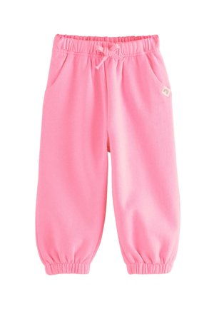 Junges Mädchen mit lockigem Haar, das einen pinken Pullover und Hose trägt, steht mit den Händen in den Taschen und bunten Sneakers auf neutralem Hintergrund.
