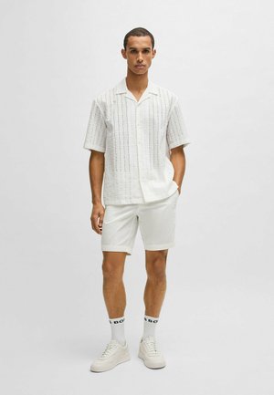 Weißes Kurzarm-Hemd mit Knöpfen und vertikalen strukturierten Streifen, kombiniert mit weißen Shorts, weißen Sneakern und weißen Socken mit schwarzen Akzenten.