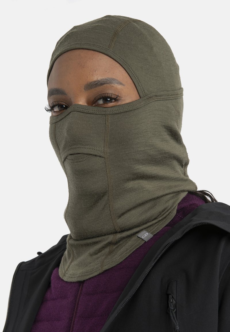 Persona que lleva un pasamontañas verde oliva con una abertura para los ojos, una camiseta morada y una chaqueta negra frente a un fondo liso.