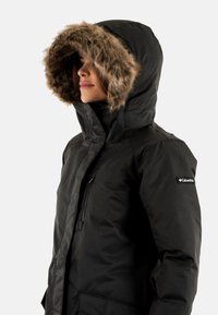 Veste d'hiver noire avec capuche à fourrure, fermeture éclair sur le devant et poches latérales. Présente un logo sur le bras gauche et une texture lisse.