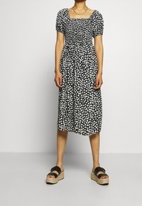 Vestido negro con estampado floral blanco, mangas abullonadas cortas, escote cuadrado y cintura fruncida. Llevado con sandalias de plataforma negras.