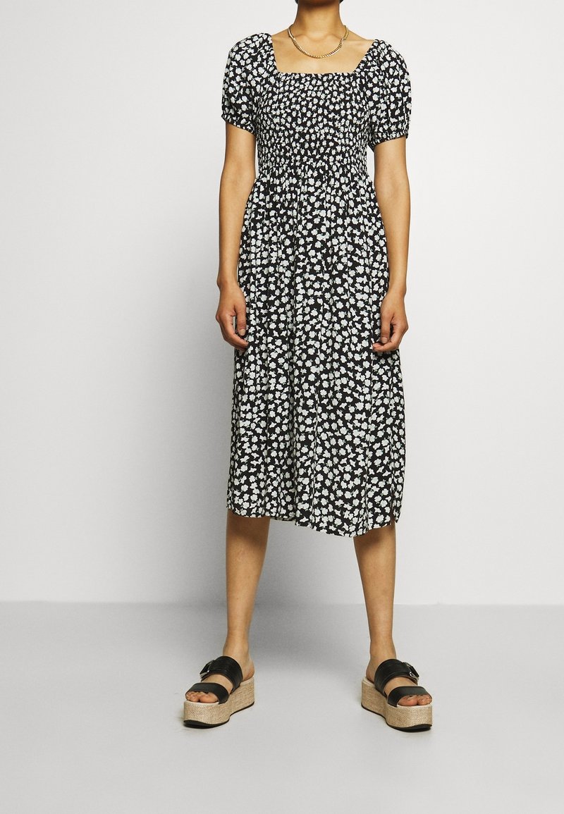 Vestido negro con estampado floral blanco, mangas abullonadas cortas, escote cuadrado y cintura fruncida. Llevado con sandalias de plataforma negras.