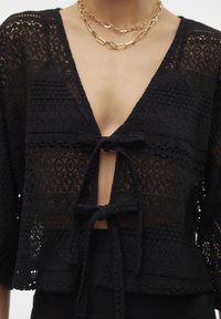 Melnas mežģīņu īsās piedurknes tops ar dziļu V-veida kaklu, sasienamu priekšpusi un sarežģītām teksturētām rakstām.