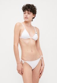 Biely bikini set obsahuje trojuholníkovú podprsenku s viazaním, textúrovanú látku a malé čierne logo na ľavej strane. Hladký povrch na nohavičkách so bočnými viazaniami.