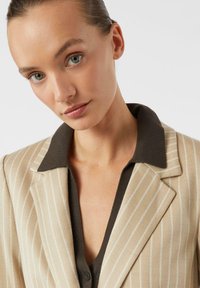Beige Nadelstreifenblazer mit braunem Reverskragen, aus strukturiertem Stoff mit betonten Schultern, vorne geknöpft.