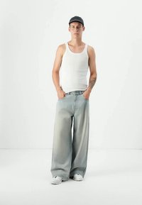 Νεαρός άνδρας φορώντας μαύρο καπέλο, λευκό αμάνικο μπλουζάκι, φαρδύ ανοιχτό μπλε τζιν και λευκά αθλητικά παπούτσια, στέκεται με τα χέρια στις τσέπες του μπροστά σε λευκό φόντο.