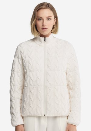 Junge Frau trägt eine weiße Strickjacke mit Reißverschluss, hohem Kragen und cremefarbene Hose vor weißem Hintergrund.