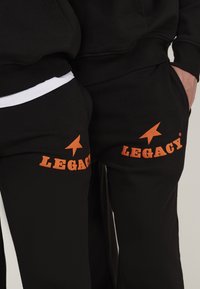 Pantalons de sport noirs avec un imprimé orange "LEGACY" et un graphique de flèche sur la cuisse, fabriqués en tissu doux avec une coupe décontractée.
