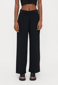 Schwarze Weitbein-Hose aus glattem Stoff mit hohem Bund und Knopfakzenten an den Seiten. Gestylt mit kurzem schwarzem Top und schwarzen Schuhen.