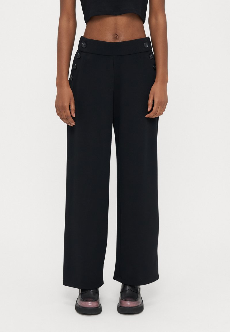 Schwarze Weitbein-Hose aus glattem Stoff mit hohem Bund und Knopfakzenten an den Seiten. Gestylt mit kurzem schwarzem Top und schwarzen Schuhen.