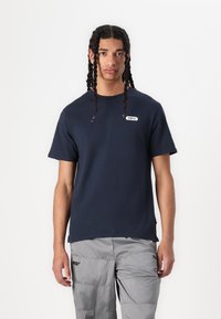 Navy short-sleeve t-shirt with a round neckline and a white label that reads "POMPEII" on the left chest, paired with gray textured pants.