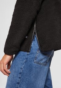 Black textured fleece jacket with side zipper partially open, worn with blue denim jeans showing back pocket and stitching details.