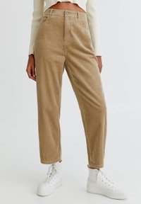 Pantalon en velours côtelé couleur beige clair, avec rayures verticales, coupe droite, taille haute et fermeture avant par boutons. Porté avec des baskets blanches.