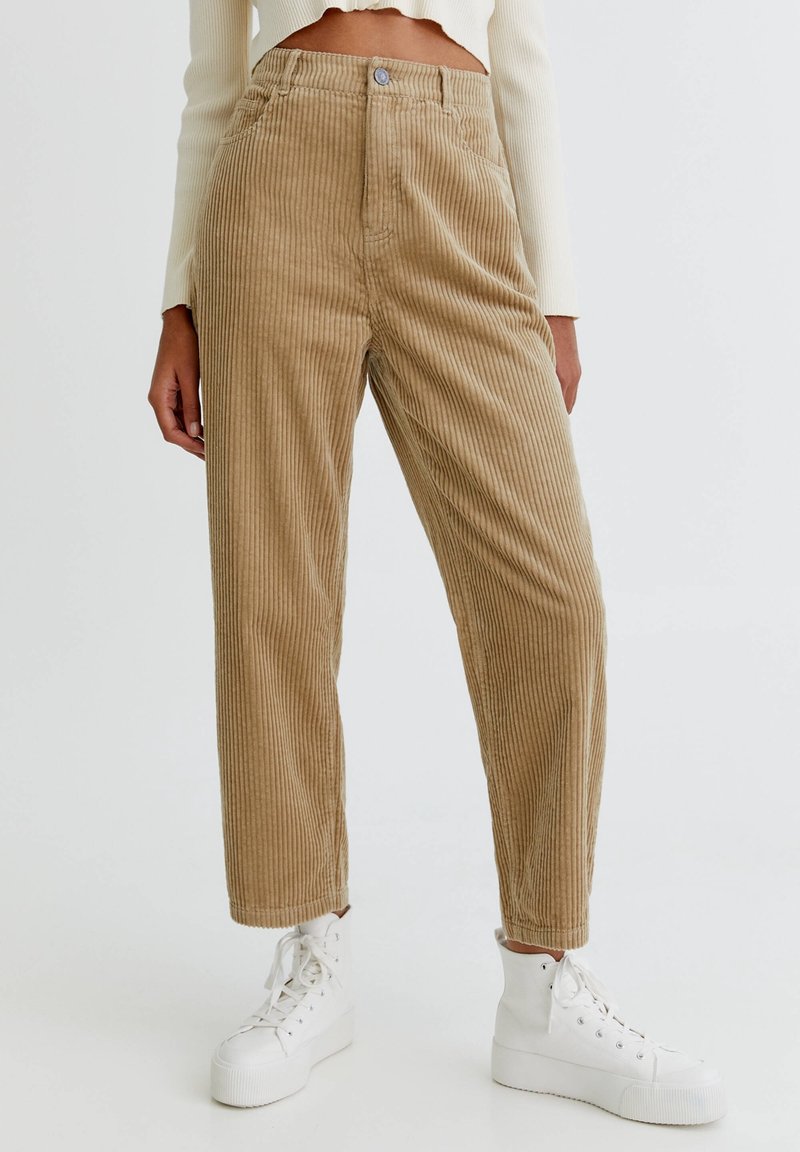 Pantalon en velours côtelé couleur beige clair, avec rayures verticales, coupe droite, taille haute et fermeture avant par boutons. Porté avec des baskets blanches.