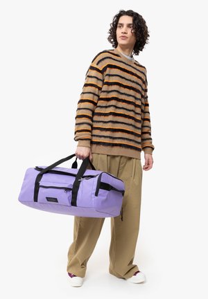 Borsa a dispenser viola con dettagli neri, dotata di manici doppi, tasca con zip e forma rettangolare. Indossata da una persona in un maglione a righe.
