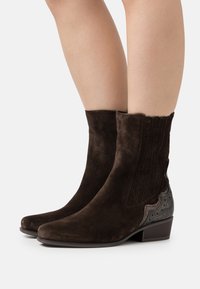 Dark brown suede ankle boots with low block heels, decorative perforations, and textured black leather accents on heels worn on bare legs.