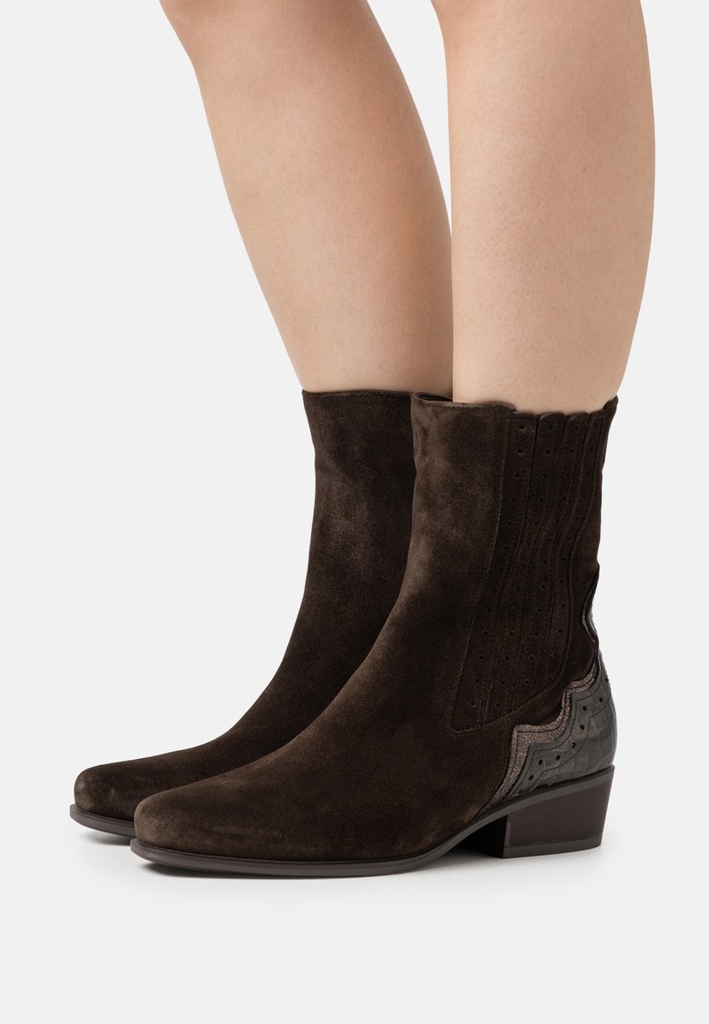 Dark brown suede ankle boots with low block heels, decorative perforations, and textured black leather accents on heels worn on bare legs.