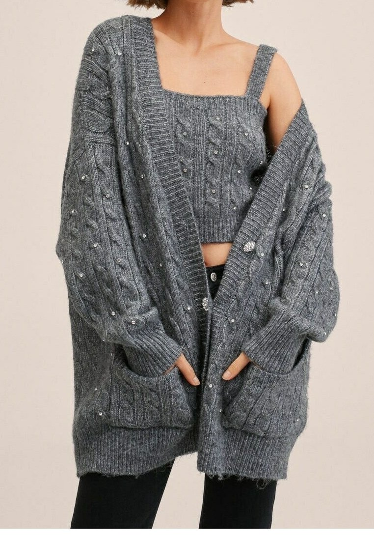 Grauer gestrickter Cardigan mit Zopfmuster, ausgestattet mit frontalen Taschen und verziert mit kleinen dekorativen Perlen. Die Ärmel sind oversized.
