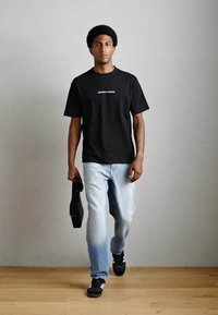 Black short-sleeve T-shirt with white text, light blue jeans, and black sneakers. Model holds a black item in the left hand. Light wood floor.
