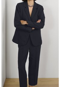 Czarnoniebieski garnitur szyty na miarę, z marynarką z klapami i prostymi spodniami. Gładki materiał o minimalistycznym wzornictwie i czystych liniach.