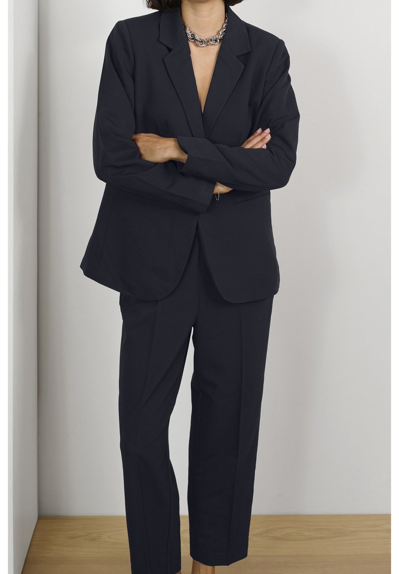 Czarnoniebieski garnitur szyty na miarę, z marynarką z klapami i prostymi spodniami. Gładki materiał o minimalistycznym wzornictwie i czystych liniach.