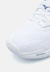 Bijela atletska cipela s teksturiranim gornjim dijelom i ojačanim potplatom. Značajke uključuju svijetloplave naglaske i "Mizuno Enerzy" brandiranje sa strane.