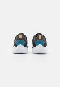 Une paire de chaussures de sport noires avec des semelles blanches, un tissu teal sur les talons et des languettes jaunes, montrée de dos sur un fond blanc.
