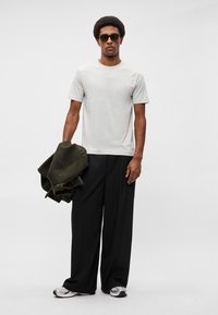 Svetloč gray tričko, široké čierne nohavice, drží olivovo zelenú bundu. Čierne slnečné okuliare, texturovaná látka, neformálny dizajn, minimálne detaily.