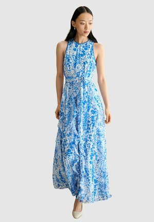 Femme portant une robe maxi sans manches à motifs floraux bleus et blancs, avec une taille cintrée et une jupe volantée, associée à des chaussures blanches fermées.