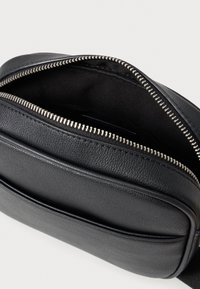 Μαύρη δερμάτινη τσάντα με υφή, που διαθέτει ασημένιο φερμουάρ και ανοιχτό πάνω μέρος που αποκαλύπτει εσωτερικό από σκούρο ύφασμα.